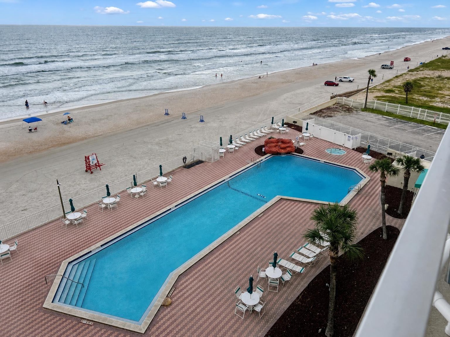 The beachfront pool stretches along a wide sandy shore where gentle waves roll in under a blue sky, offering direct access to the ocean from your vacation retreat.