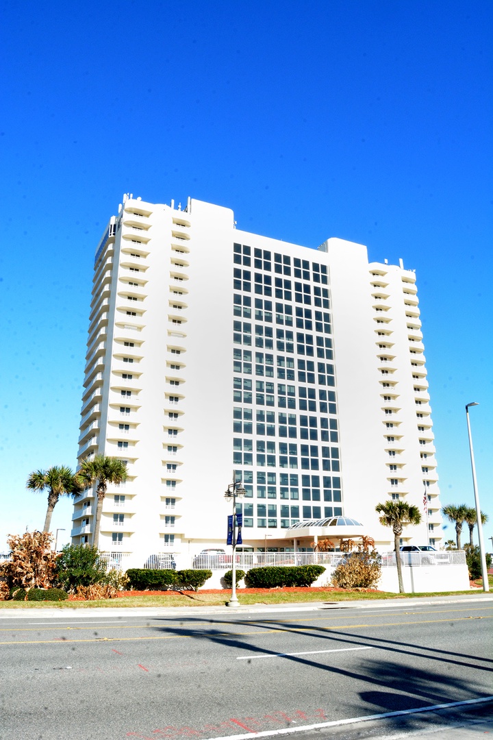 Modern high-rise property featuring distinctive white architecture with palm-lined entrance, set against brilliant blue skies in a prime location.
