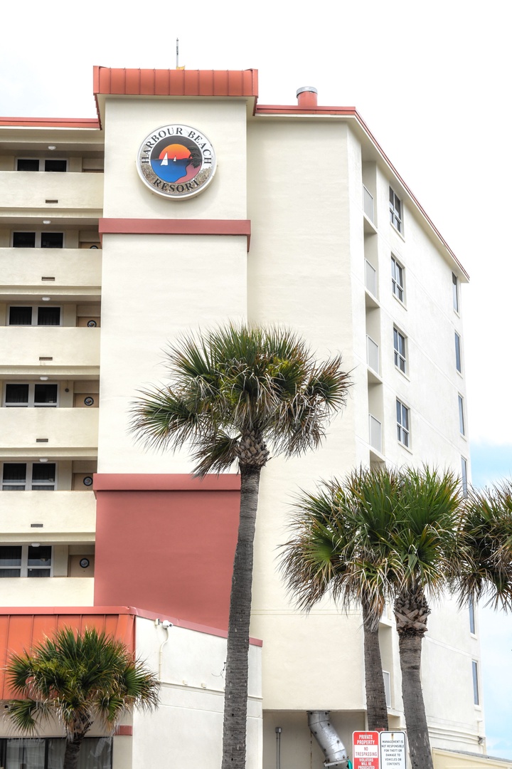 A mid-rise beachfront property with distinctive coral and cream architecture, framed by swaying palm trees under a coastal sky.