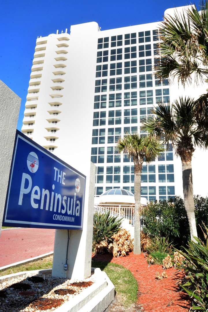 Modern white condominium building with blue Peninsula sign and tropical palm landscaping under clear skies.