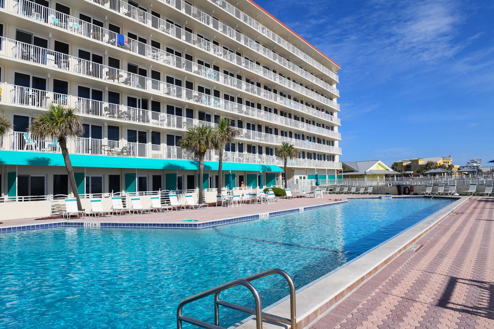 A beachside resort building with turquoise awnings overlooks a sparkling outdoor pool, surrounded by sun loungers and palm trees under clear skies.