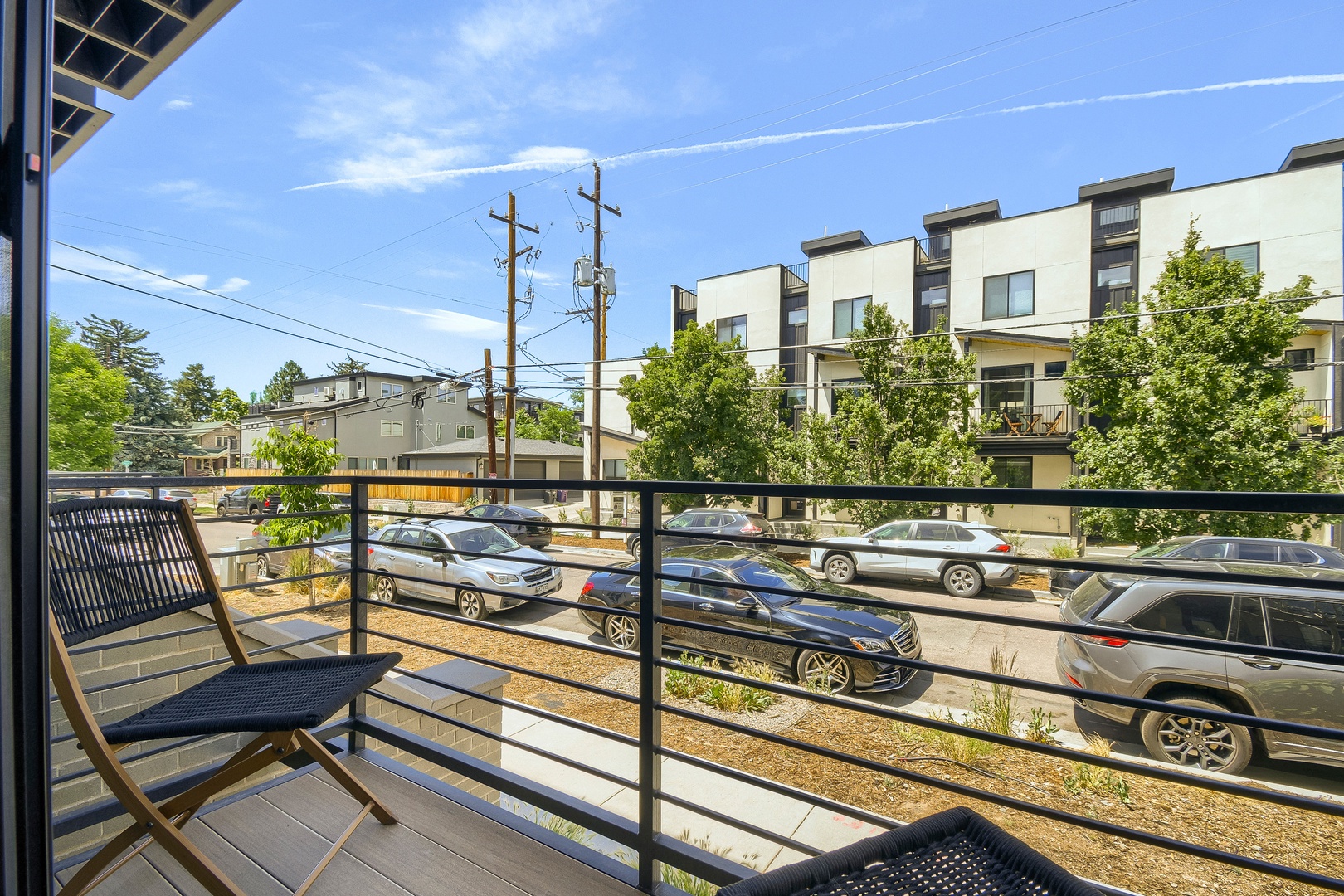 Modern outdoor balcony with sleek metal railings and comfortable seating overlooks the surrounding neighborhood on a bright, sunny day.