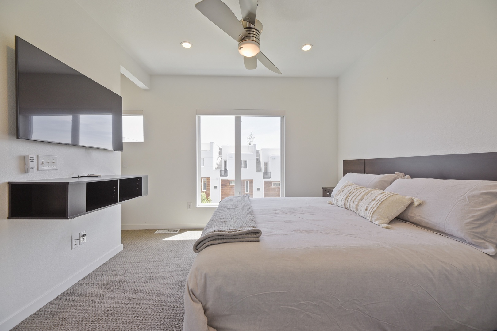 Your modern retreat awaits with a plush bed, ceiling fan overhead, wall-mounted TV, and abundant natural light streaming through large windows—perfect for restful mornings and peaceful evenings.