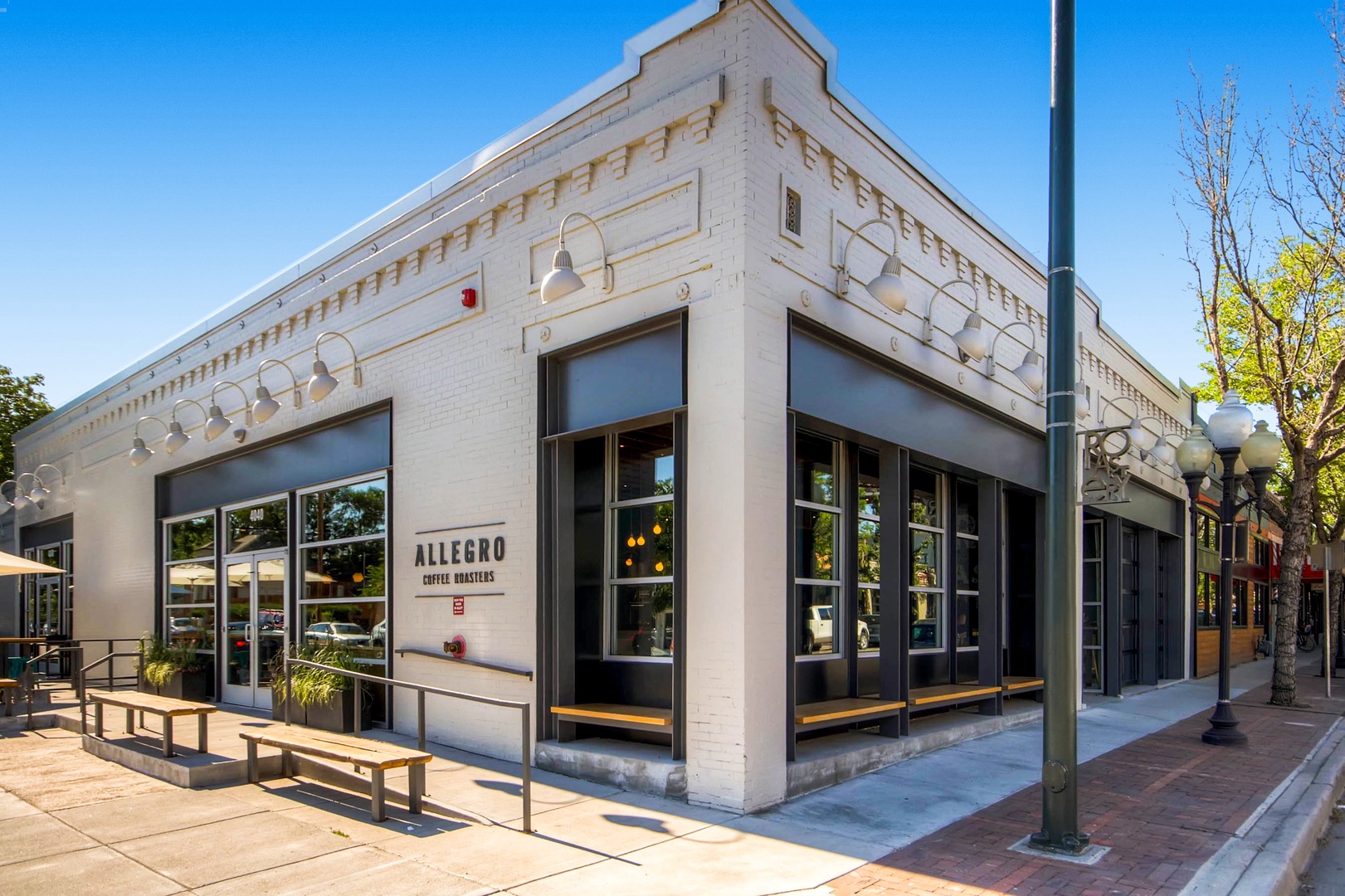 A white-painted corner building with large windows and outdoor seating marks the entrance to Allegro Coffee Roasters, located in a walkable neighborhood setting along a tree-lined street.