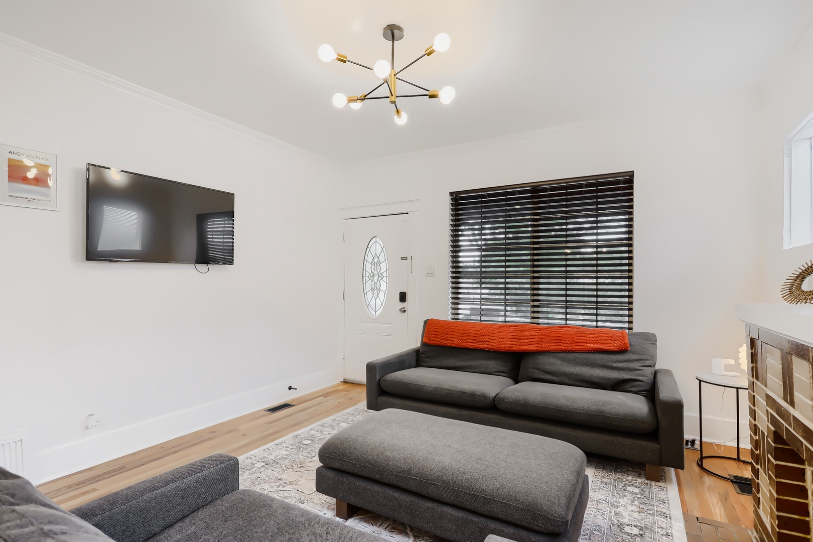 Step into your stylish living room where modern design meets relaxation. Unwind on the plush gray sofa under warm lighting, catch your favorite shows on the flat-screen, and enjoy the natural light streaming through horizontal blinds.