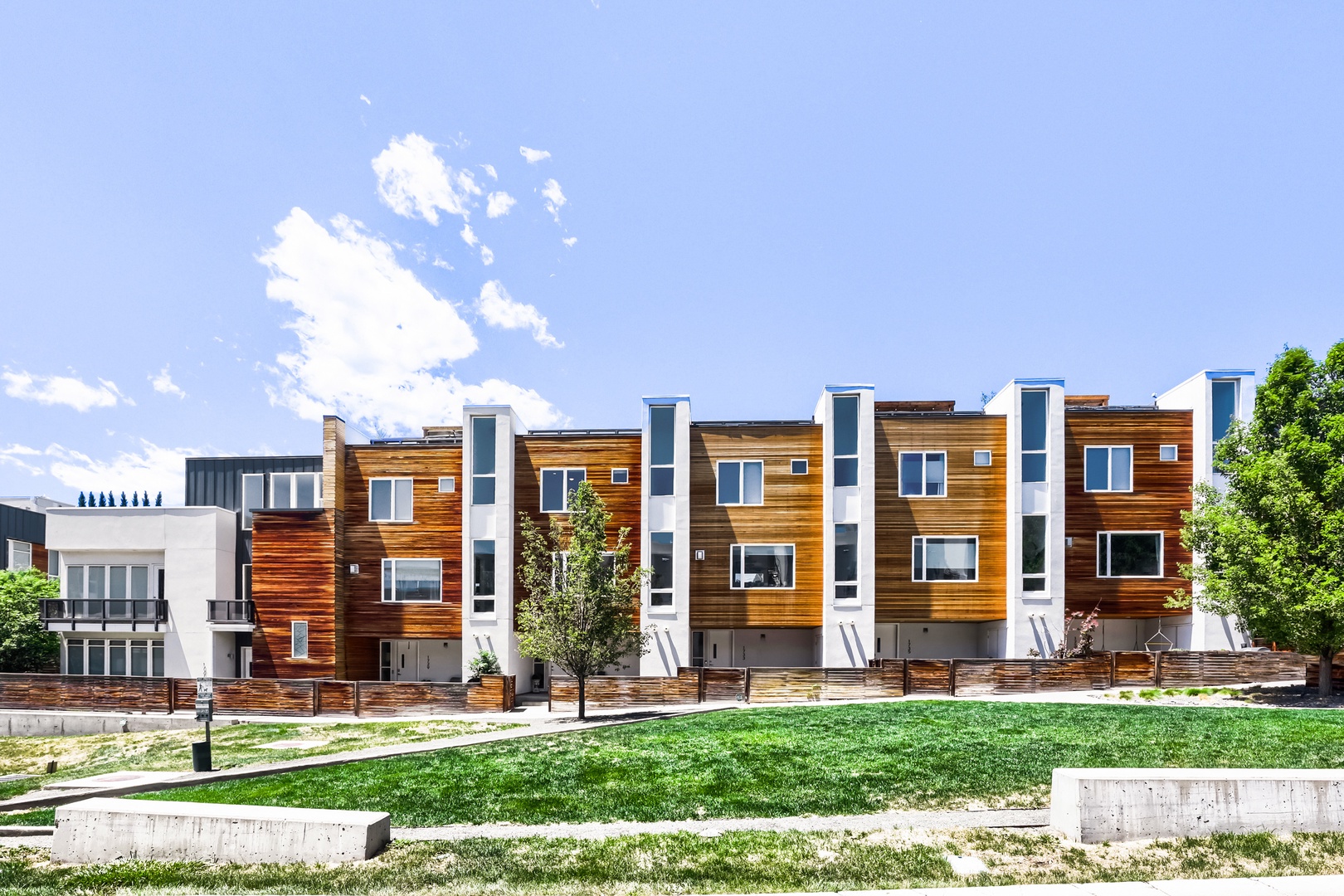 Modern townhome community features warm wood-clad exteriors with white accents and private ground-floor entrances. A well-maintained green lawn and neighboring properties visible in the surrounding residential area.