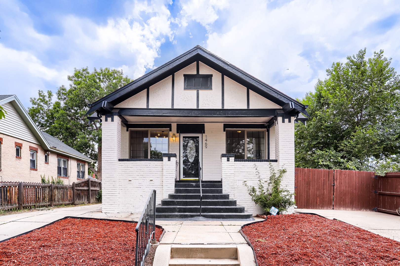 A charming craftsman-style bungalow with distinctive dark trim and white brick exterior, nestled in a tree-lined residential neighborhood. The property features a welcoming front porch and well-maintained landscaping with red mulch beds.