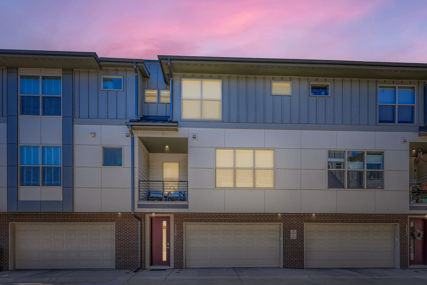 Modern townhome exterior featuring warm lighting and contemporary architecture against a colorful dusk sky. The attached garage provides convenient parking at your arrival.