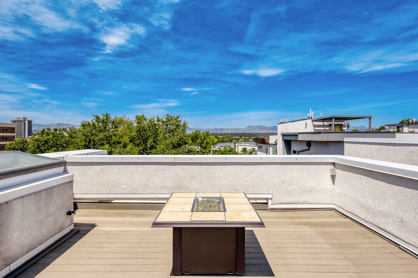 Modern rooftop terrace featuring a central fire table, spacious deck, and panoramic views of the surrounding neighborhood, mountains, and lush greenery.