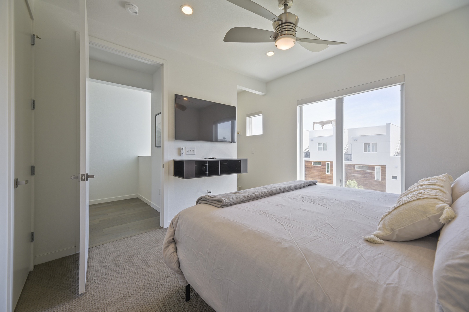 Wake up to natural light streaming through large windows in this serene bedroom. Ceiling fan keeps you comfortable while the sleek wall-mounted TV and modern furnishings create your perfect retreat.