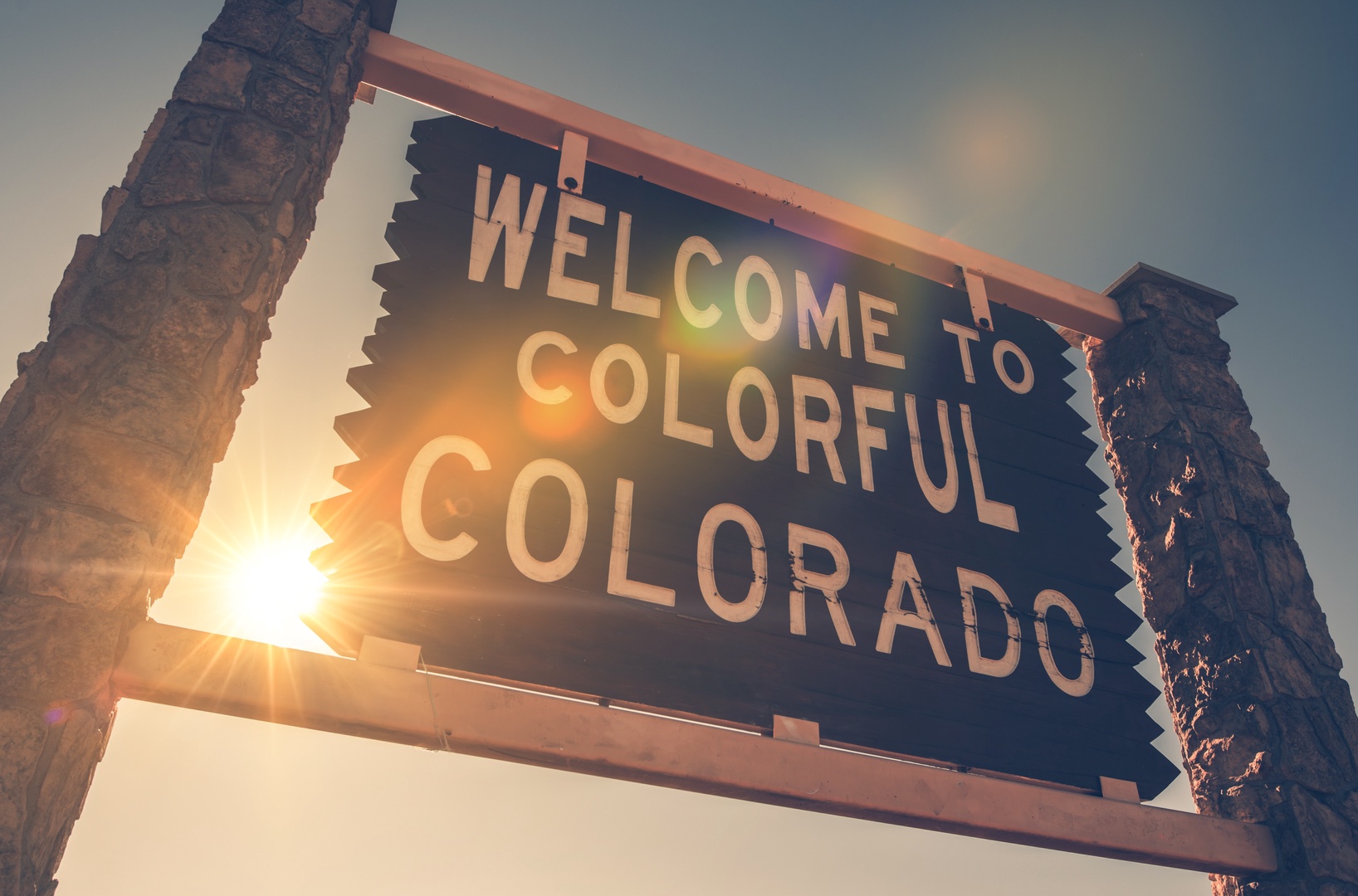 The iconic "Welcome to Colorful Colorado" sign marks the entrance to the state, framed by sunlit stone pillars as a bright beacon of arrival.