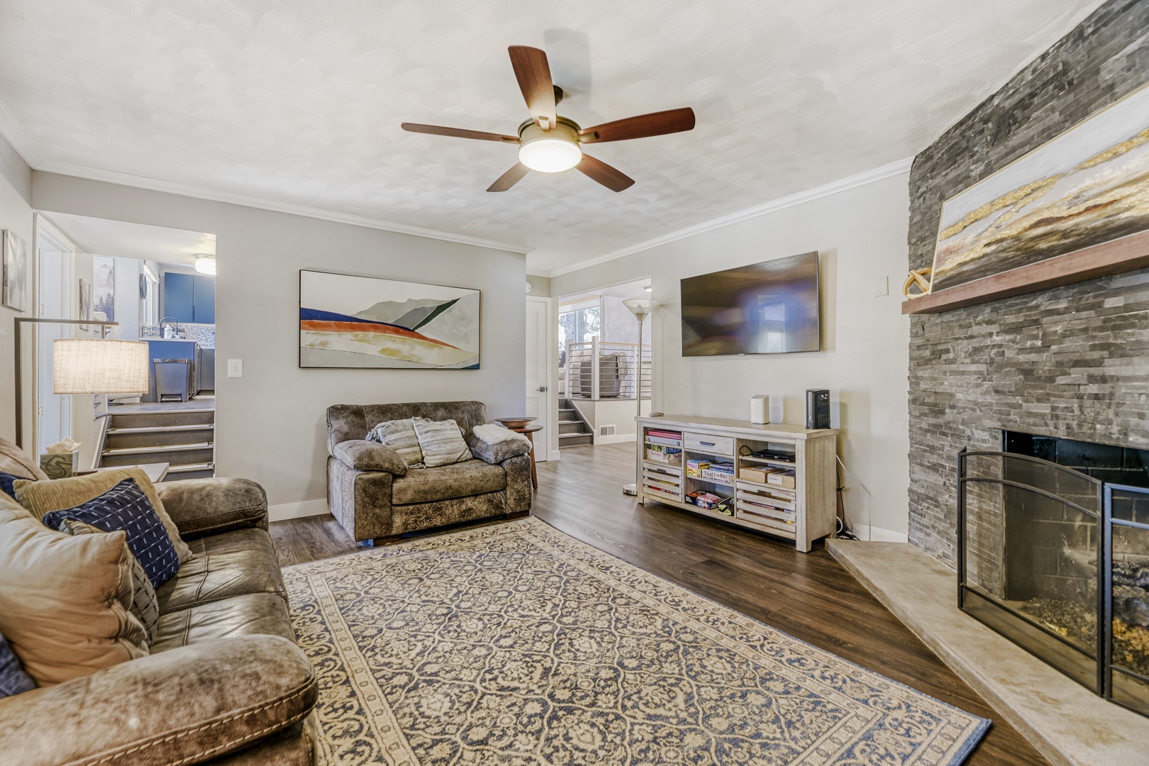 Your living room invites you to unwind with plush seating, warm wood accents, and a striking stone fireplace. Modern entertainment awaits under soft ceiling fan breezes.