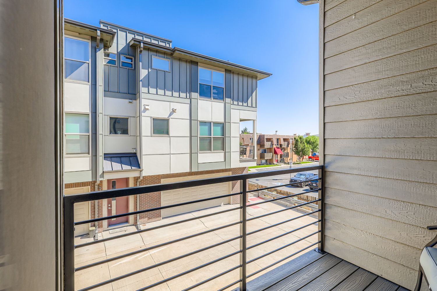 A private balcony offers views of the modern neighborhood and nearby residential buildings under bright blue skies.