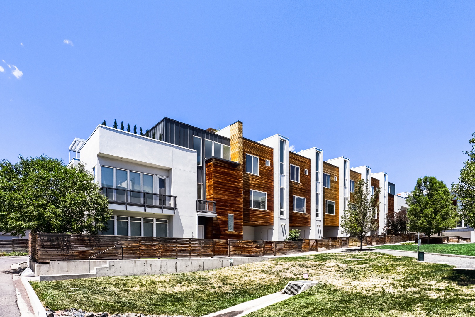 Modern multi-unit property featuring contemporary architecture with wood and white paneling, situated in a residential neighborhood with mature trees and open green spaces.