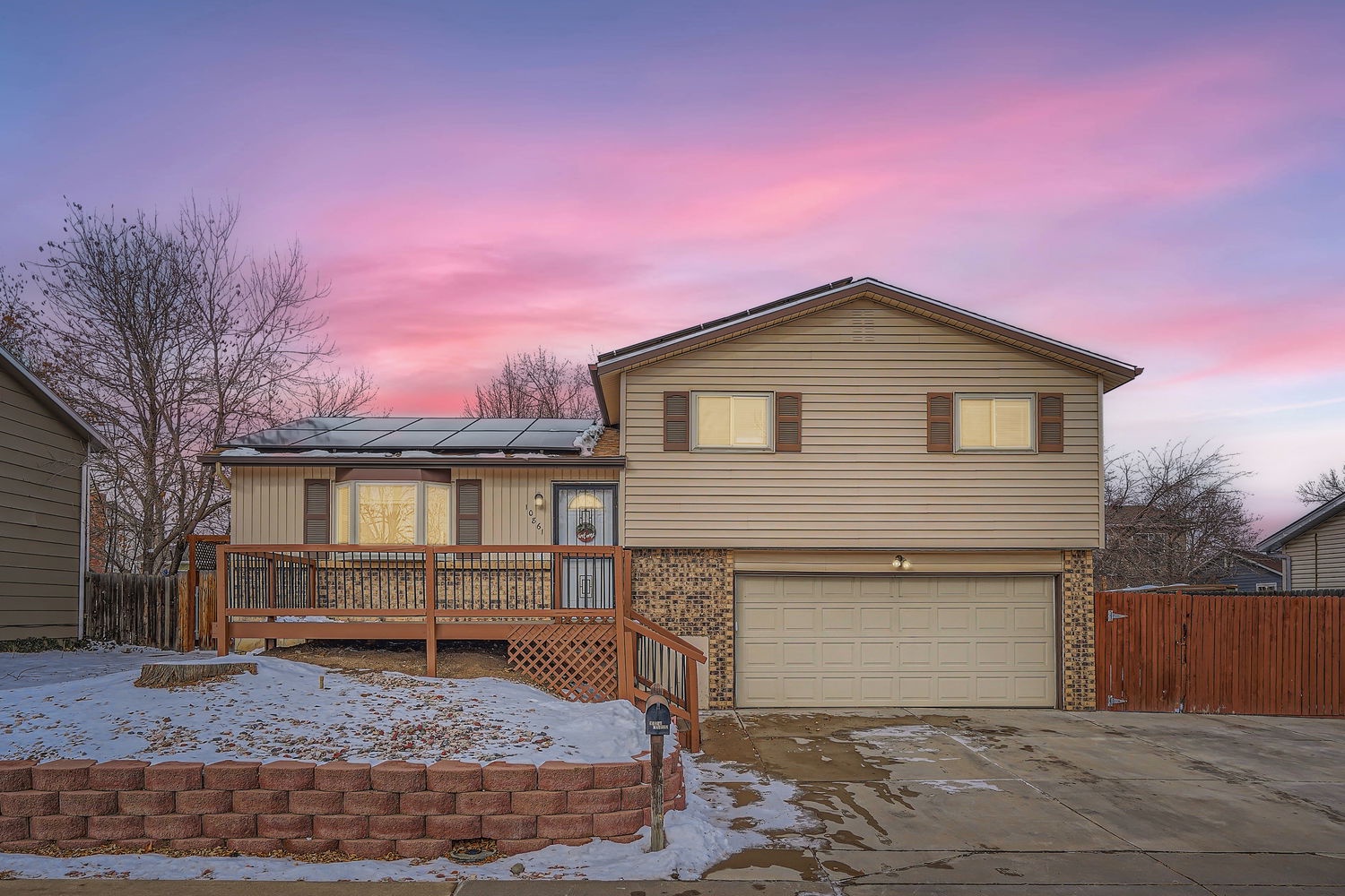 A two-story home with warm lighting and a welcoming front deck, set against a stunning winter sunset. Convenient parking and a peaceful neighborhood setting surround this property.