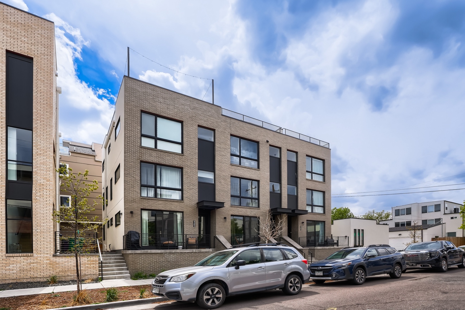 Modern multi-unit residential building in a quiet neighborhood setting, featuring contemporary brick and dark panel architecture with street parking available.