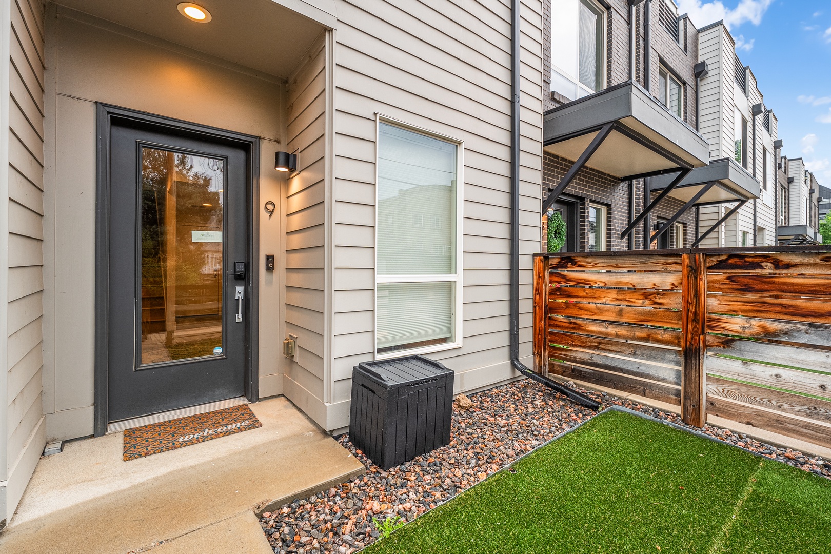 Modern townhouse entrance with private front patio featuring a covered entryway, charming wooden fence, and low-maintenance landscaping—perfect for your arrival and relaxing outdoors.