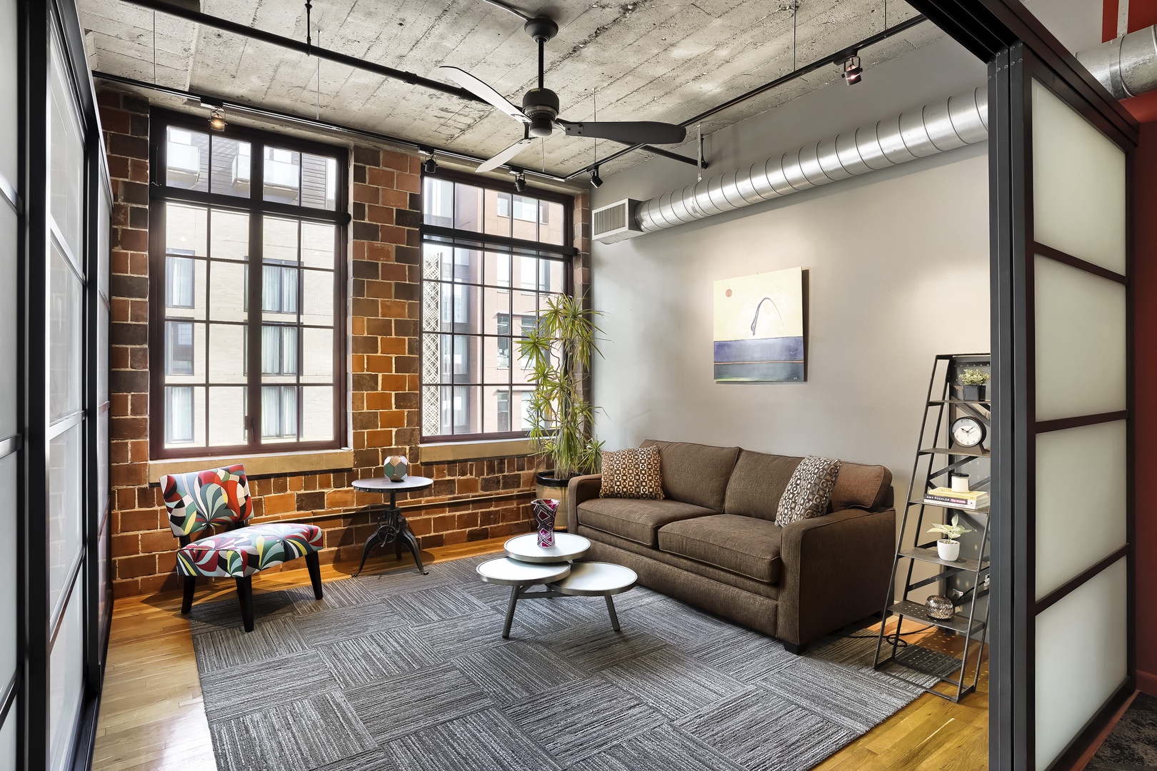 Step into this urban loft where exposed brick meets industrial charm. Sink into the plush sofa, bathed in natural light from floor-to-ceiling windows, and let the warm hardwood and high ceilings create your perfect city retreat.