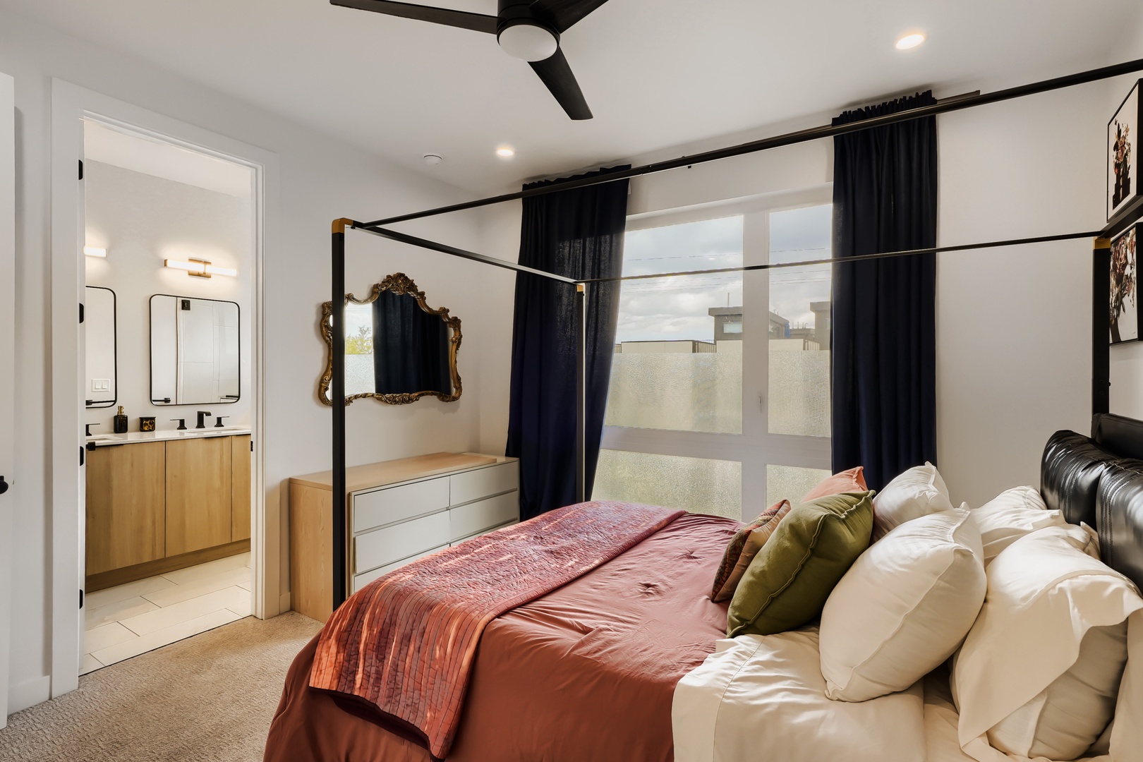 Sink into the luxurious four-poster bed, framed by elegant black drapes and an ornate mirror. Modern comforts blend with warm design touches, while natural light floods through generous windows. Your private en-suite awaits just beyond.