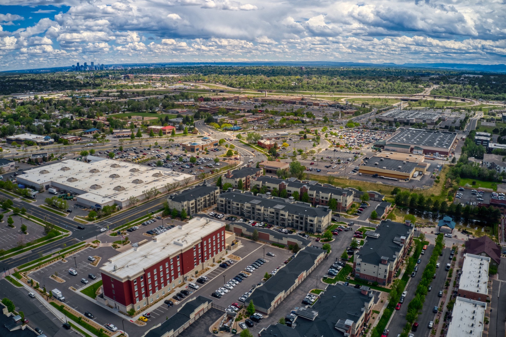 Aerial perspective of the property amid a mixed-use neighborhood with abundant trees, commercial centers, and easy highway access. The distant city skyline marks the metropolitan area beyond.