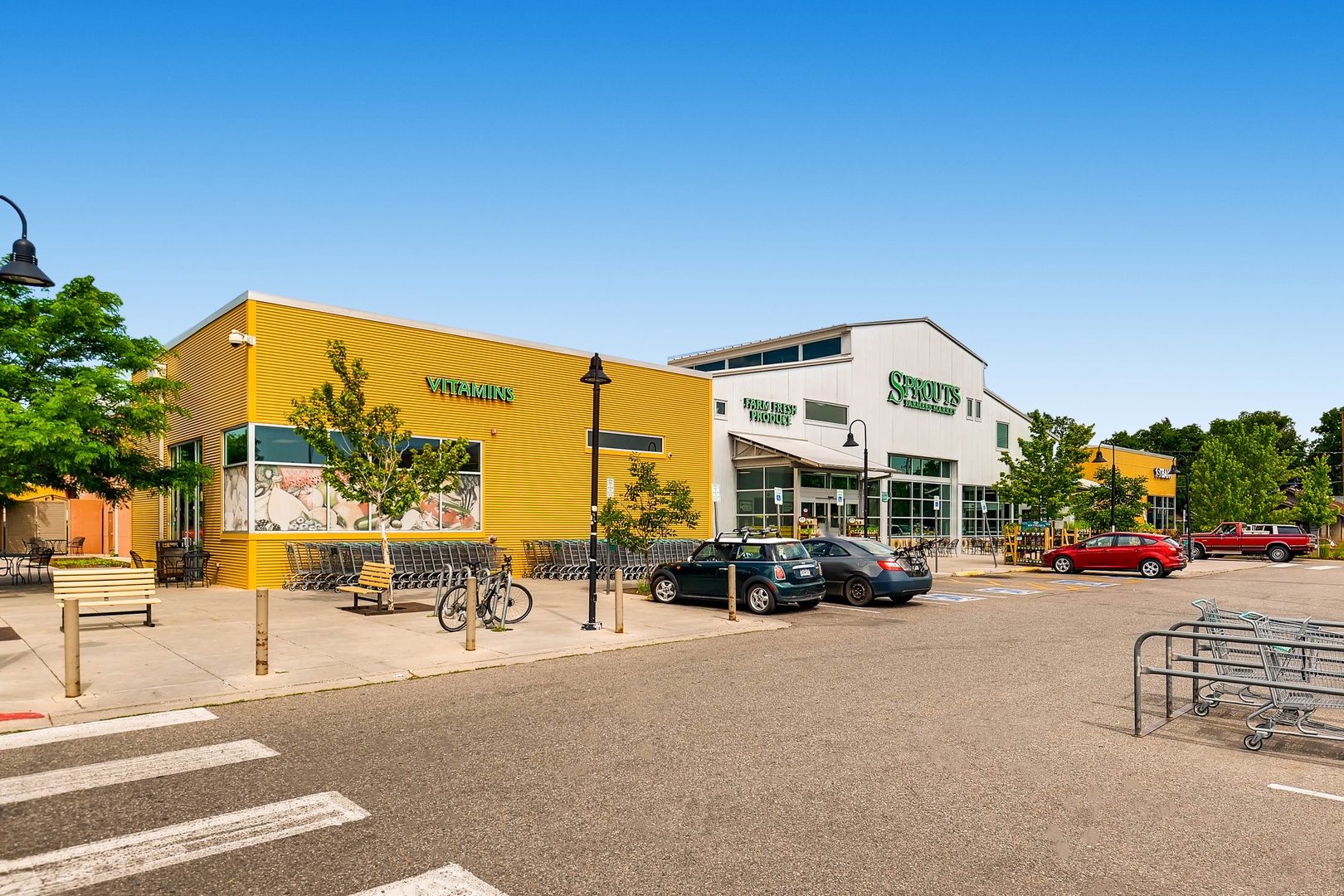 Vibrant local shopping area features a bright yellow Vitalkins building and Sprout's Farmers Market, both set in a modern retail plaza surrounded by lush greenery and ample parking.