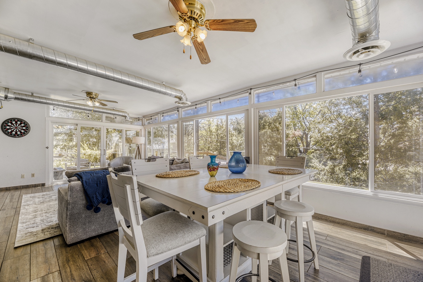 Your sun-drenched dining space awaits, where walls of windows frame lush treetop views. Gather at the crisp white table for memorable meals, cooled by gentle ceiling fans above.