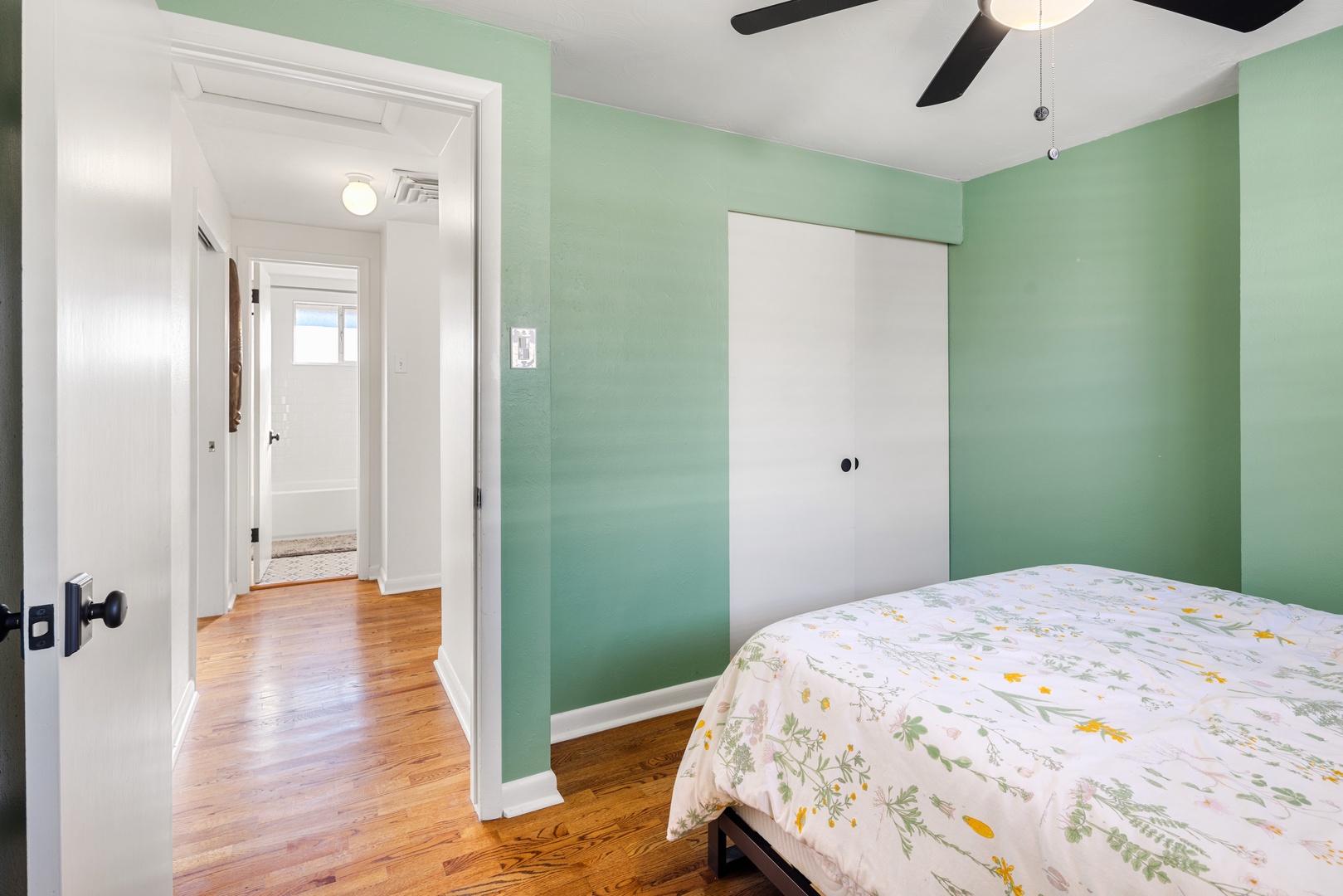 Unwind in your tranquil green bedroom where floral bedding and warm hardwood floors create a peaceful retreat after exploring the city.