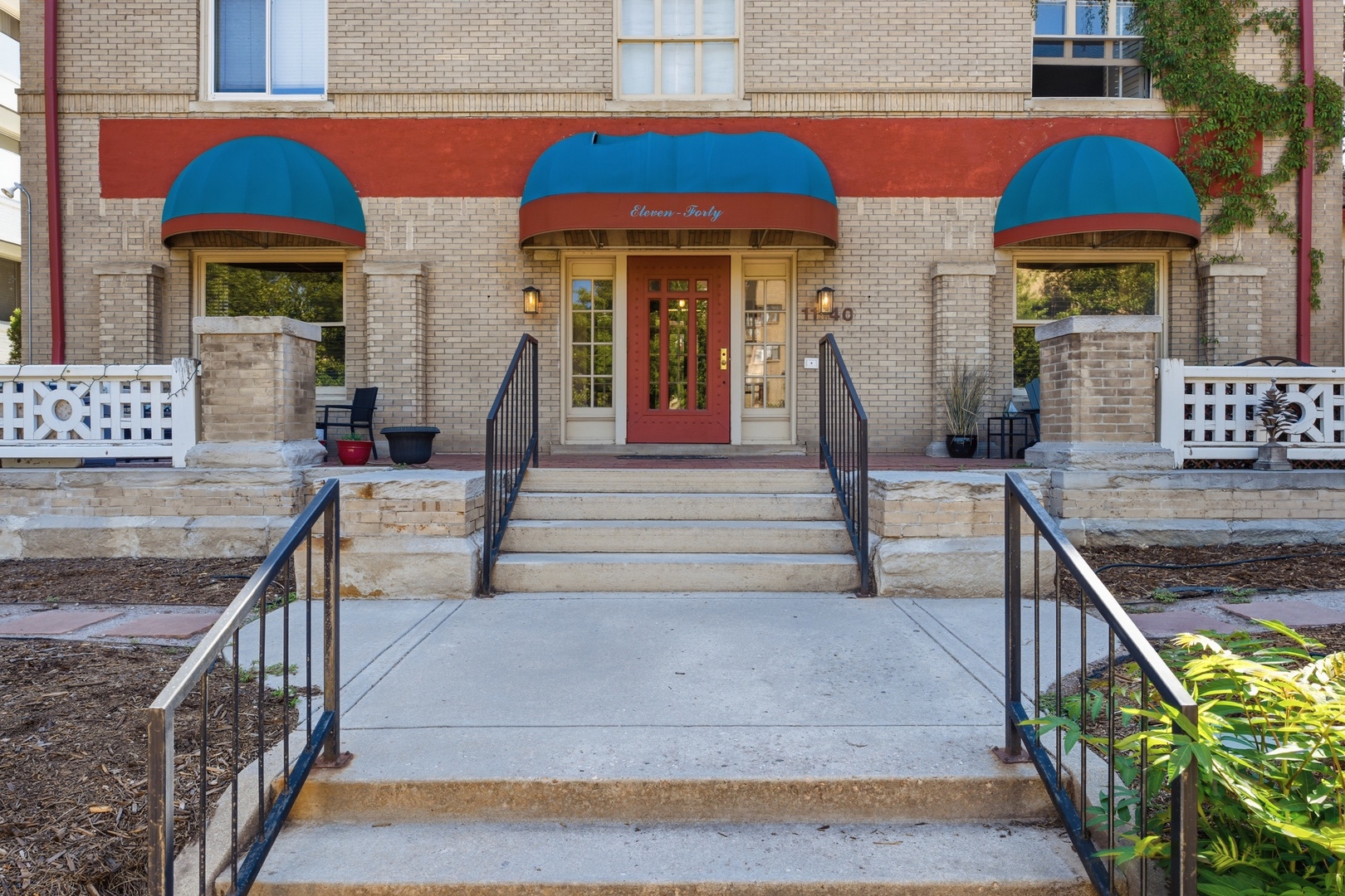 The property's welcoming facade features charming red and blue awnings framing the main entrance, with accessible ramps and seating areas flanking stone steps.