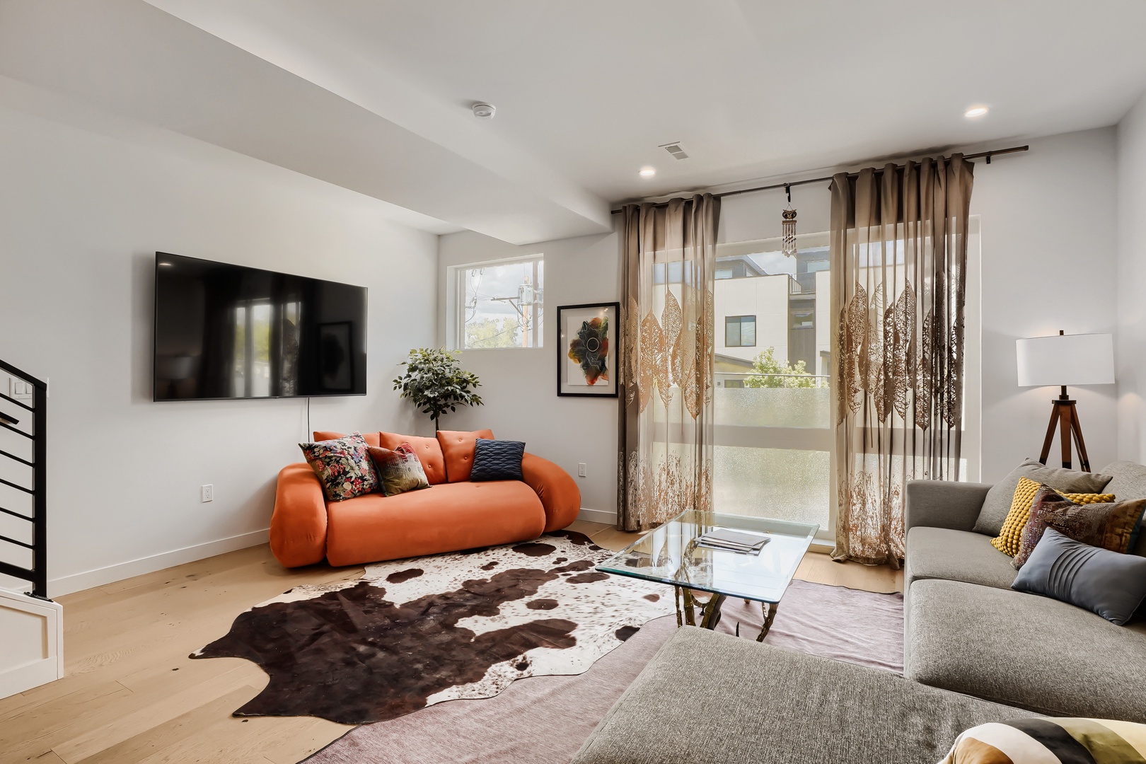 Unwind in your bright, modern living space with a vibrant orange sofa and stylish cowhide rug. Natural light pours through sheer curtains as you relax in this inviting retreat.