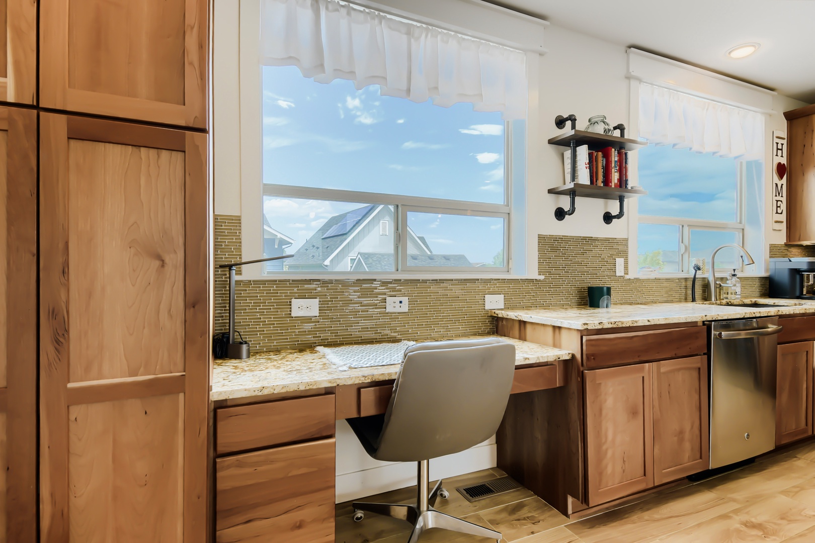 Imagine cooking in this bright kitchen with its warm wood cabinetry, granite counters, and built-in desk—all bathed in natural light from windows framing blue skies.