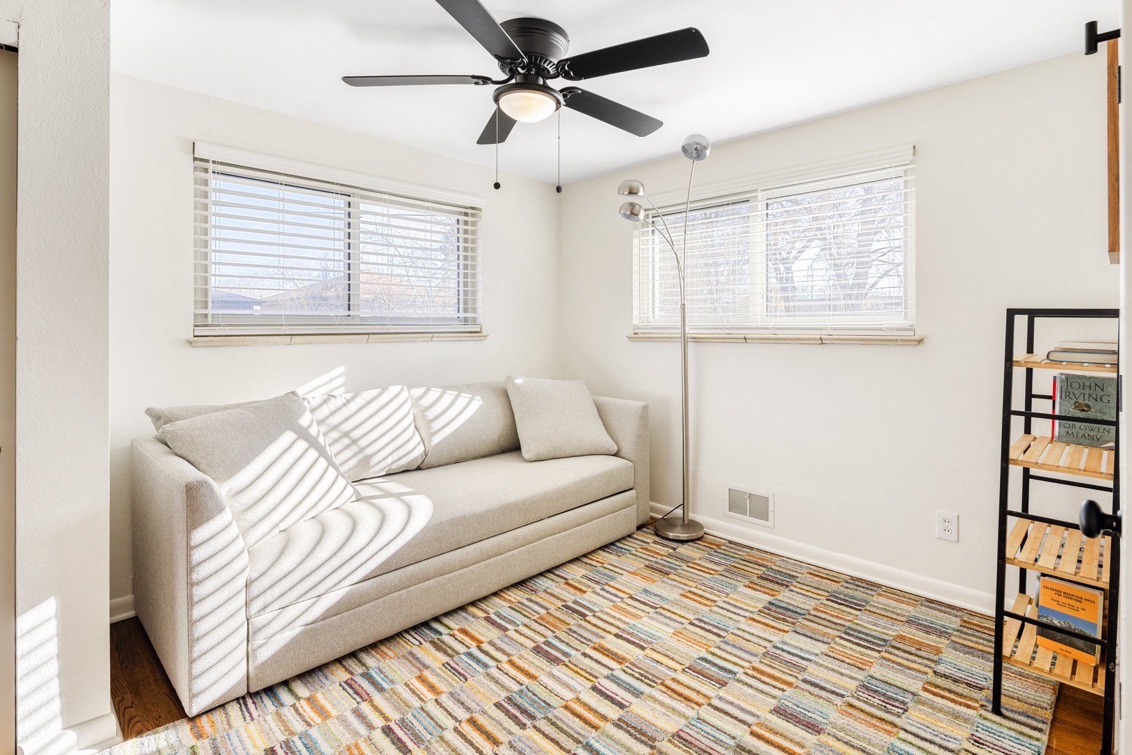 Relax in your bright, airy living space where natural light streams through windows onto the comfortable sofa. The ceiling fan keeps you cool.