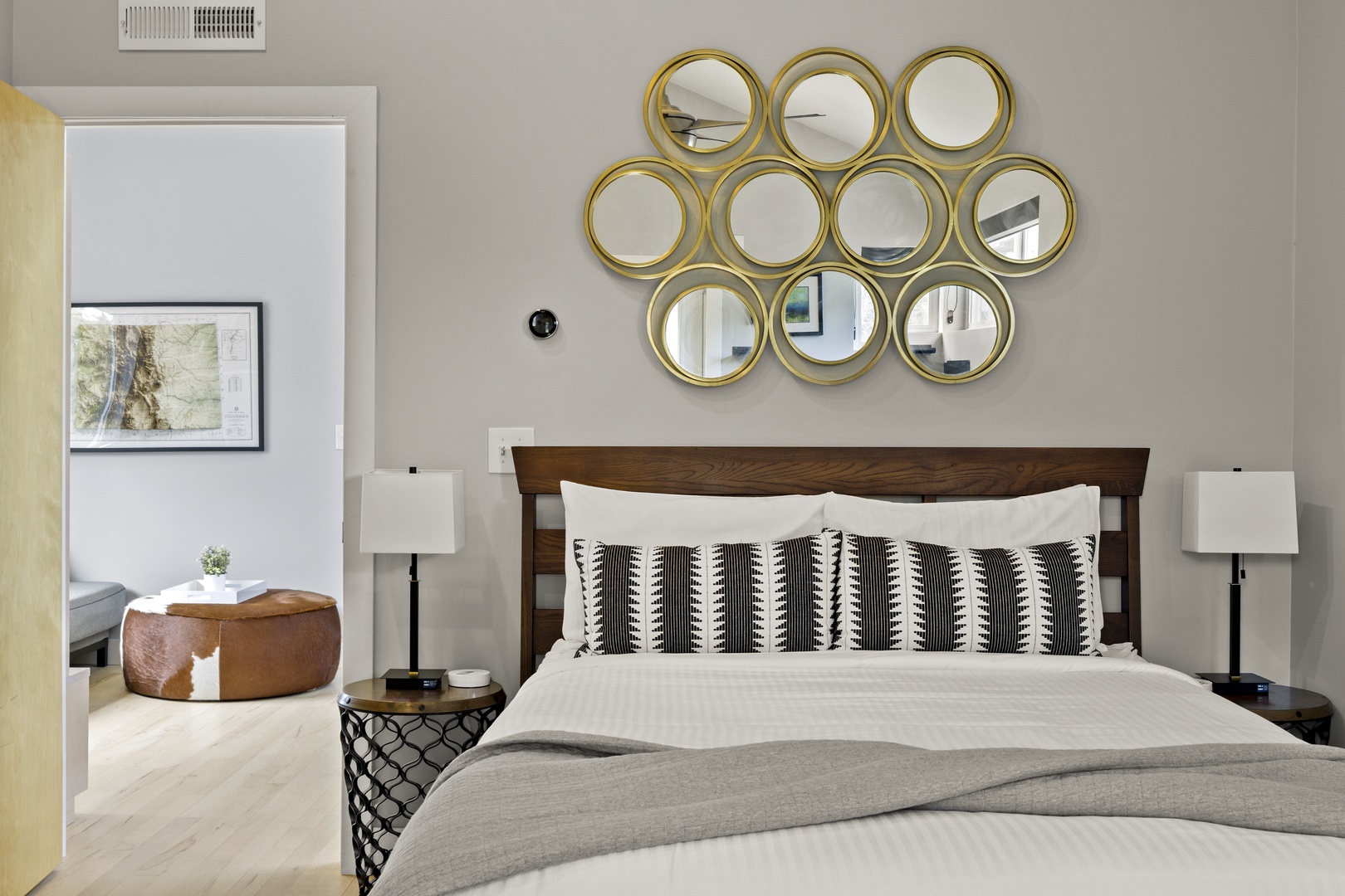 Rest peacefully in your stylish bedroom featuring a plush bed, striking gold mirror wall art, and cozy seating area visible through the doorway.