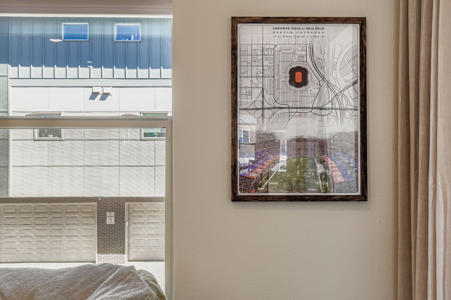 A framed map of Empower Field at Mile High and its surroundings adds local character to this bright bedroom, while the window offers views of the building across the street in this Denver location.