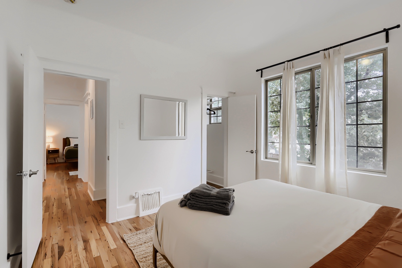Bright, minimalist bedroom with natural light streaming through large windows. The warm hardwood floors and crisp linens create a peaceful retreat where you can unwind and recharge.