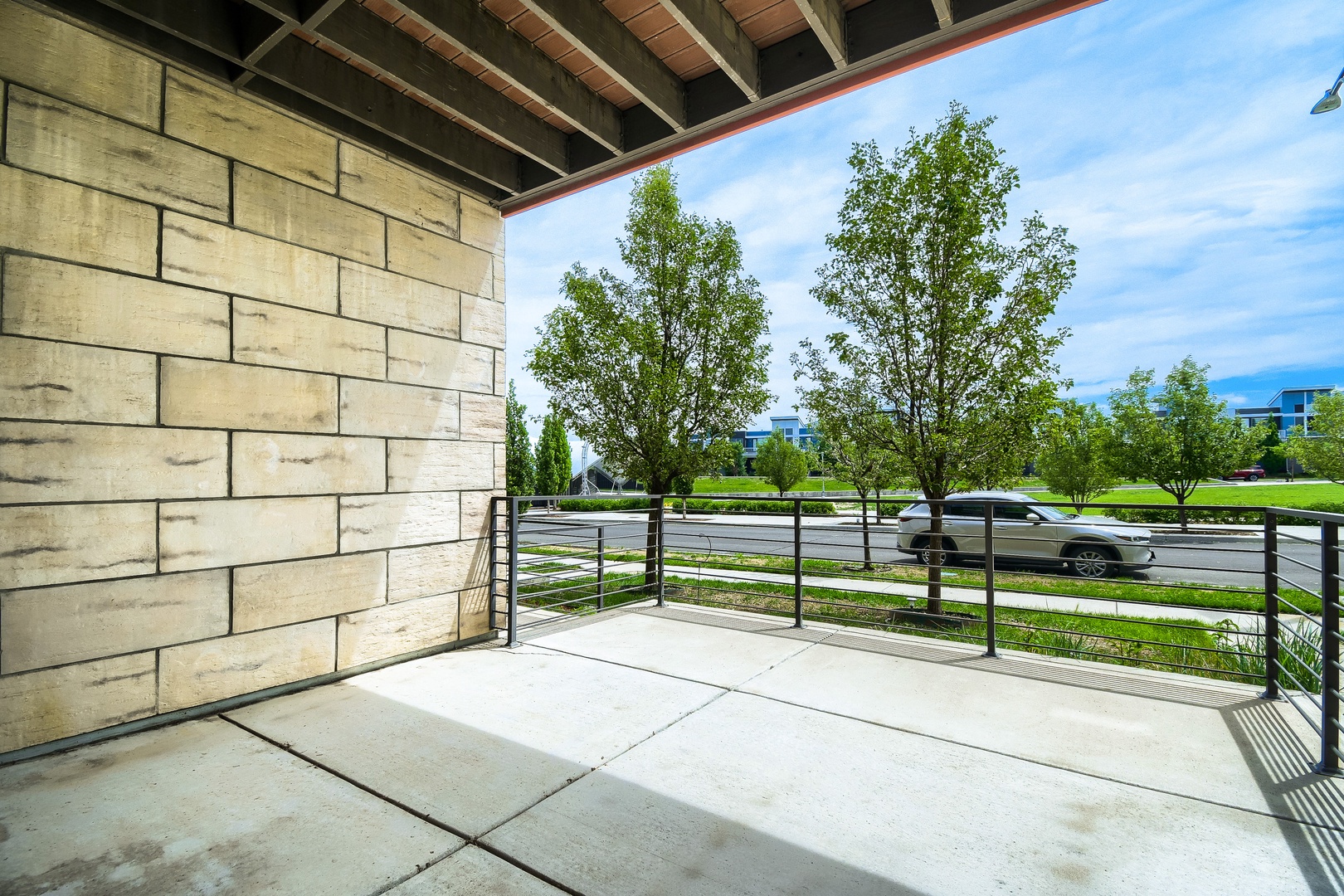 A covered patio with modern metal railing overlooks a tree-lined street and nearby greenspace. The property features easy street access and parking convenience.