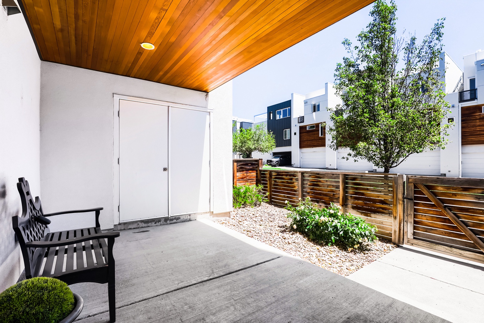 Modern covered patio with wood-slat ceiling, featuring a private courtyard with mature trees and contemporary fencing. Adjacent to neighborhood with modern multi-level homes.