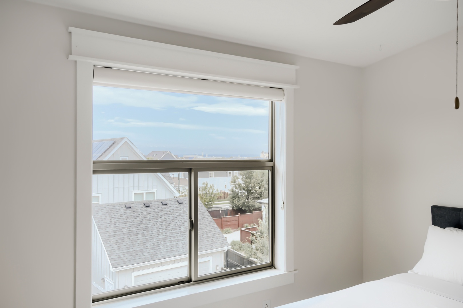 Your bedroom window frames a charming neighborhood view of coastal rooftops and distant ocean horizon, bathed in soft natural light from the ceiling fan above.