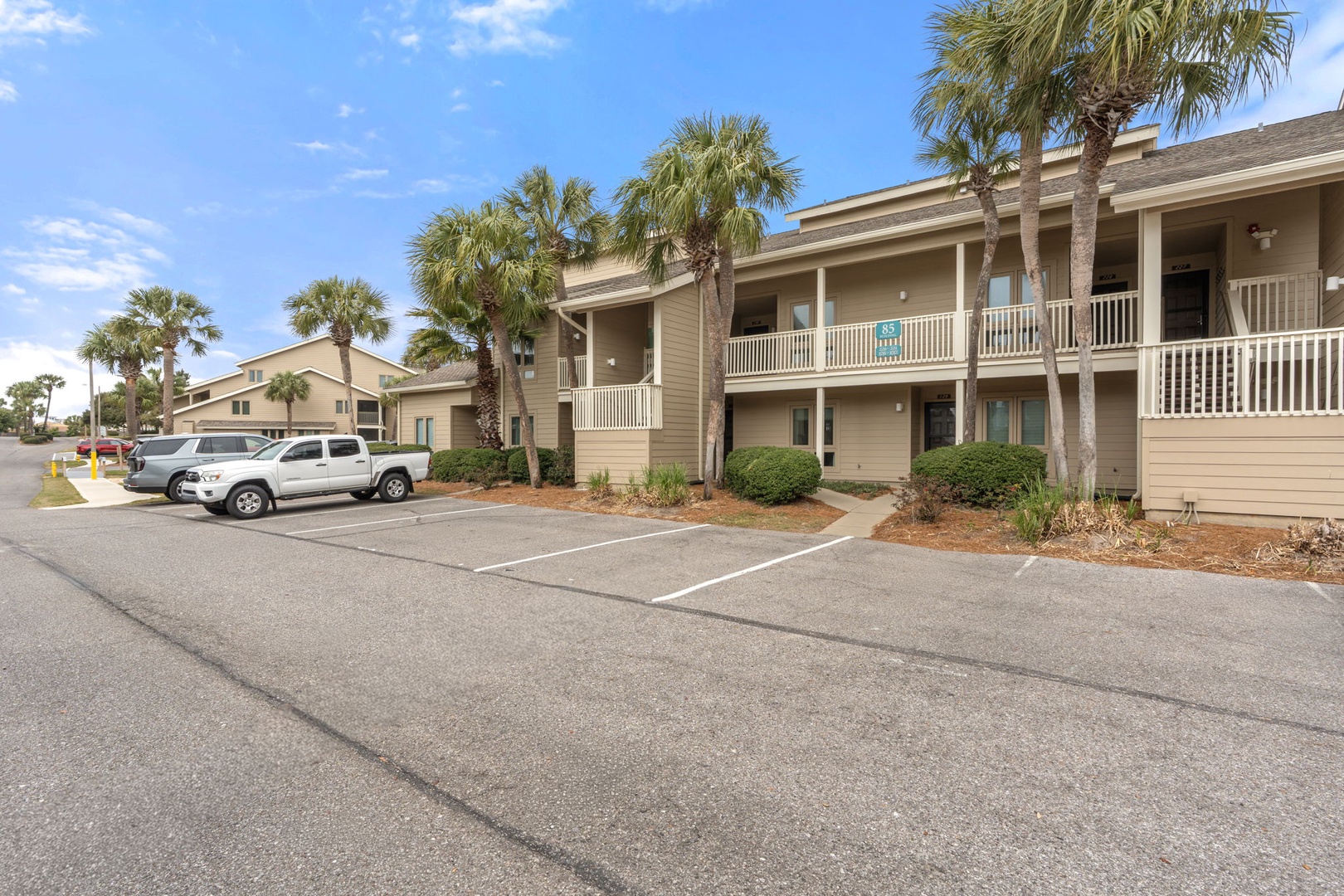 Two-story beige vacation rental building features white balcony railings and palm trees, with convenient parking spaces available for guests.