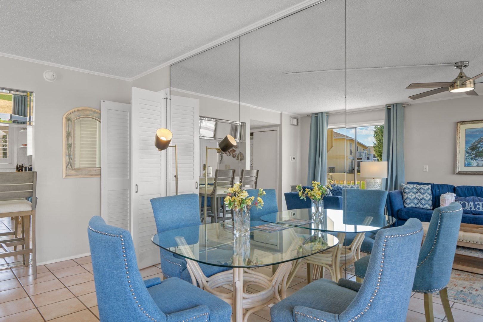 The dining area features a glass-top table with seating for four, coastal-inspired décor, and an open layout that flows seamlessly into the living space. A bright and inviting spot for meals, morning coffee, or gathering after a day at the beach.
