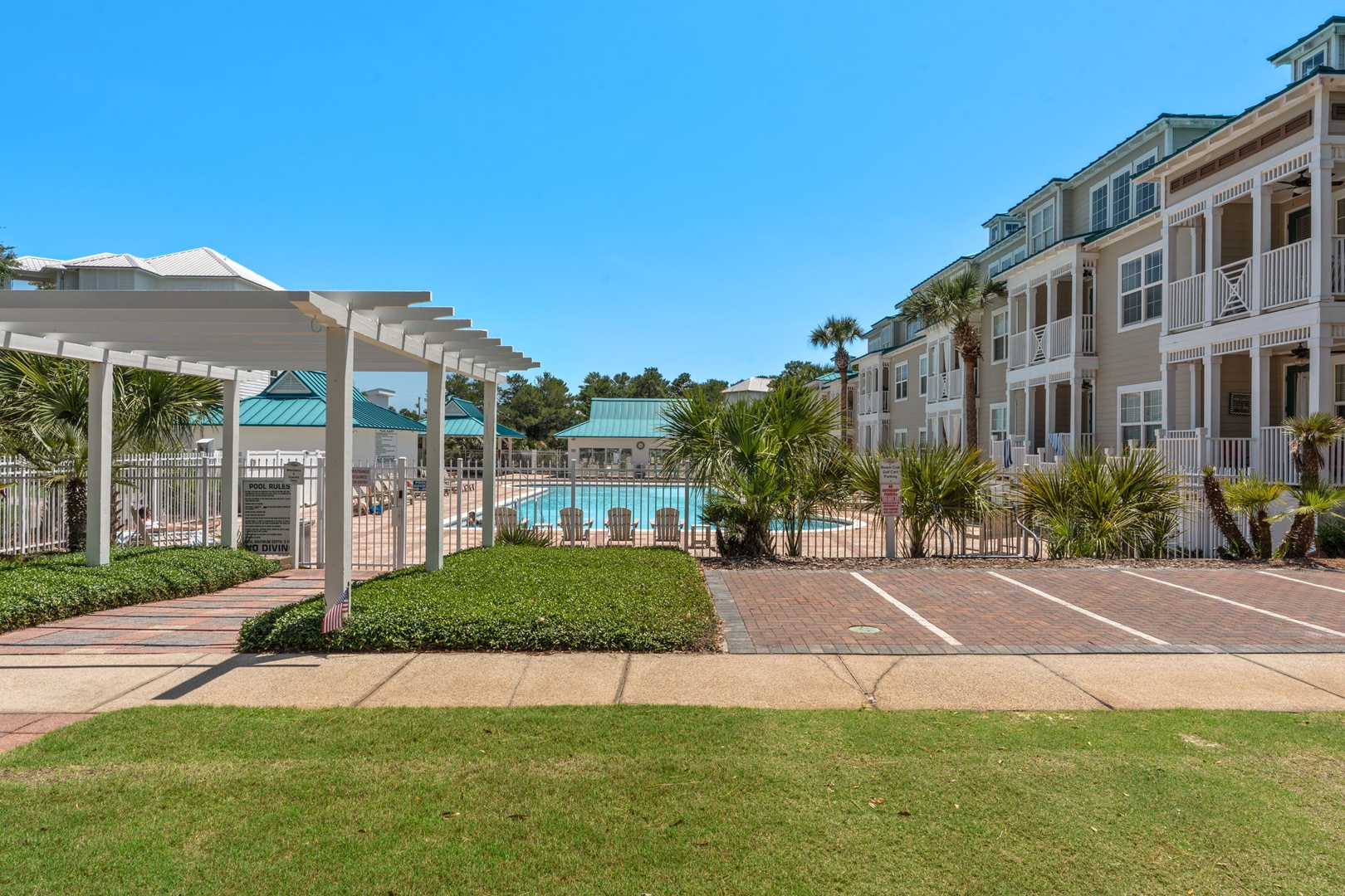 The property features a sparkling swimming pool surrounded by manicured landscaping, palm trees, and multi-story buildings with charming balconies. A white pergola and paved walkways complete this inviting coastal retreat setting.