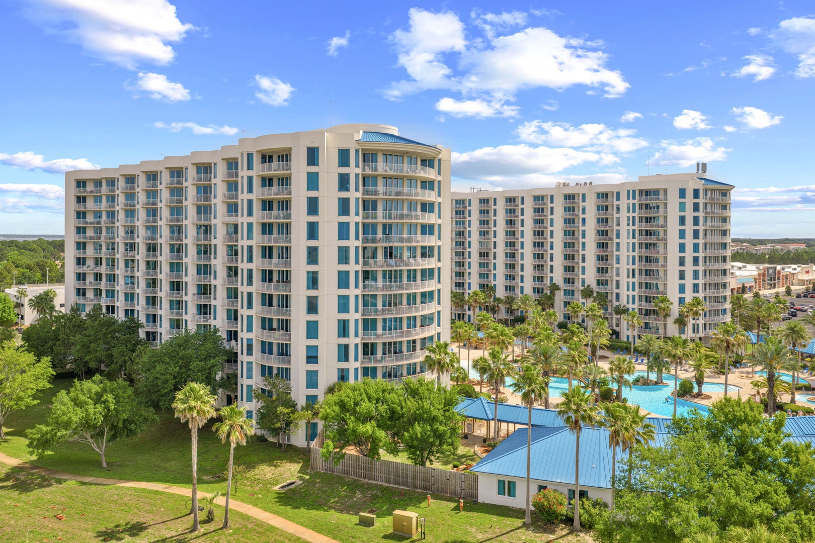 A modern multi-story resort property surrounded by lush landscaping, palm trees, and green spaces under a bright blue sky.