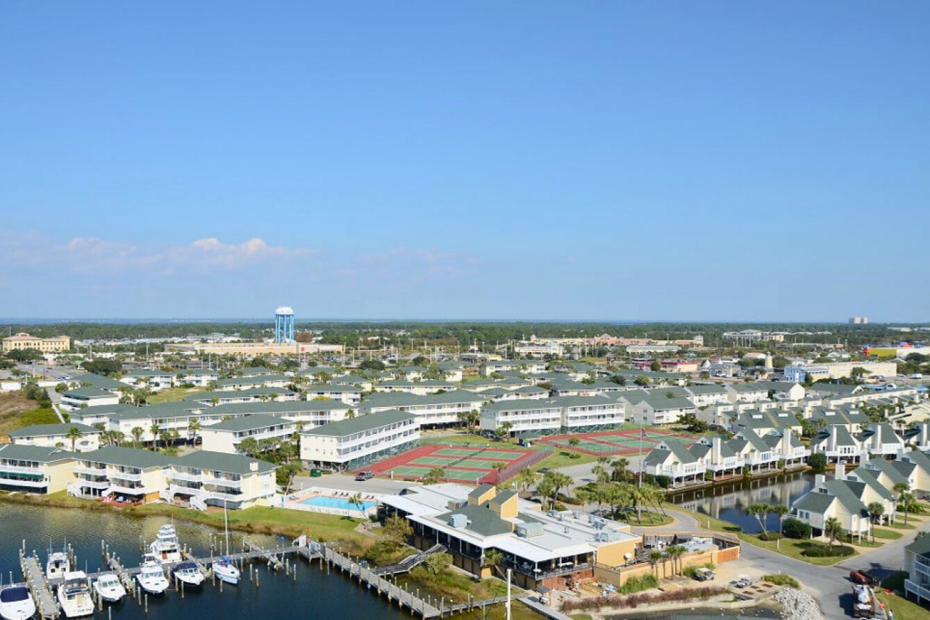 Aerial view of waterfront resort complex with marina, tennis courts, pool facilities and nearby residential community.