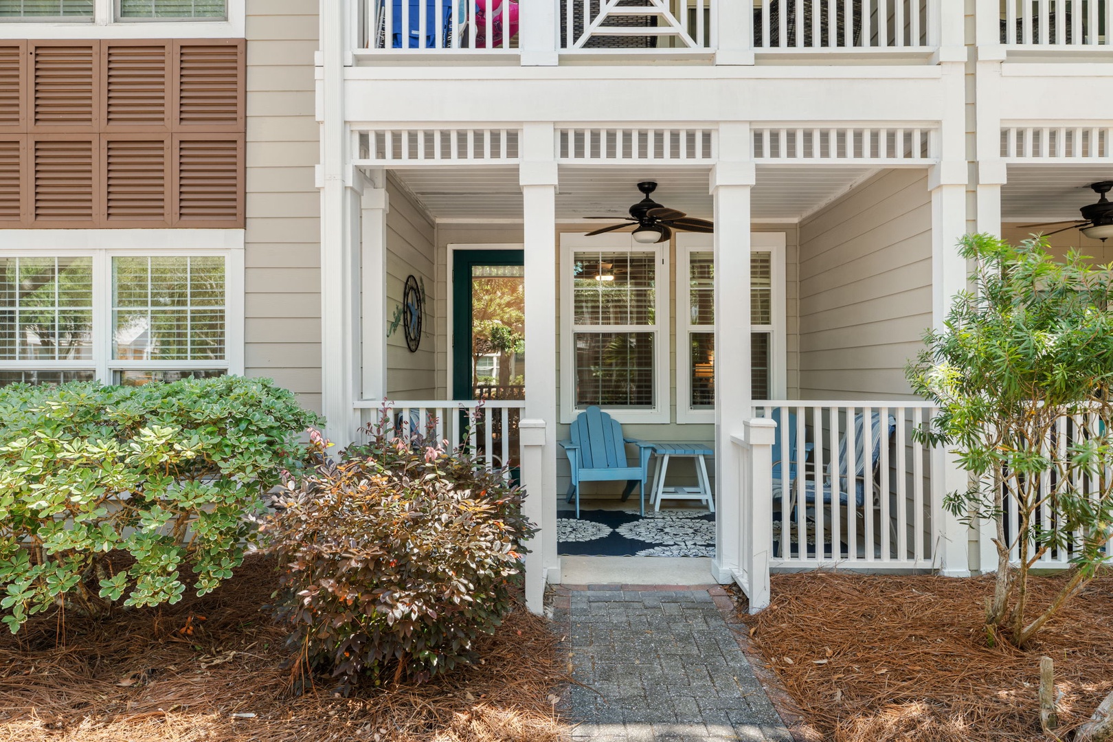 A charming coastal-style entrance welcomes you with classic white railings, a covered porch with ceiling fan, and lush greenery framing the pathway to your vacation retreat.