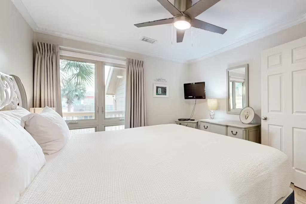 Relax in this comfortable bedroom featuring a plush bed, wall-mounted TV, ceiling fan, and large windows that bring in plenty of natural light.