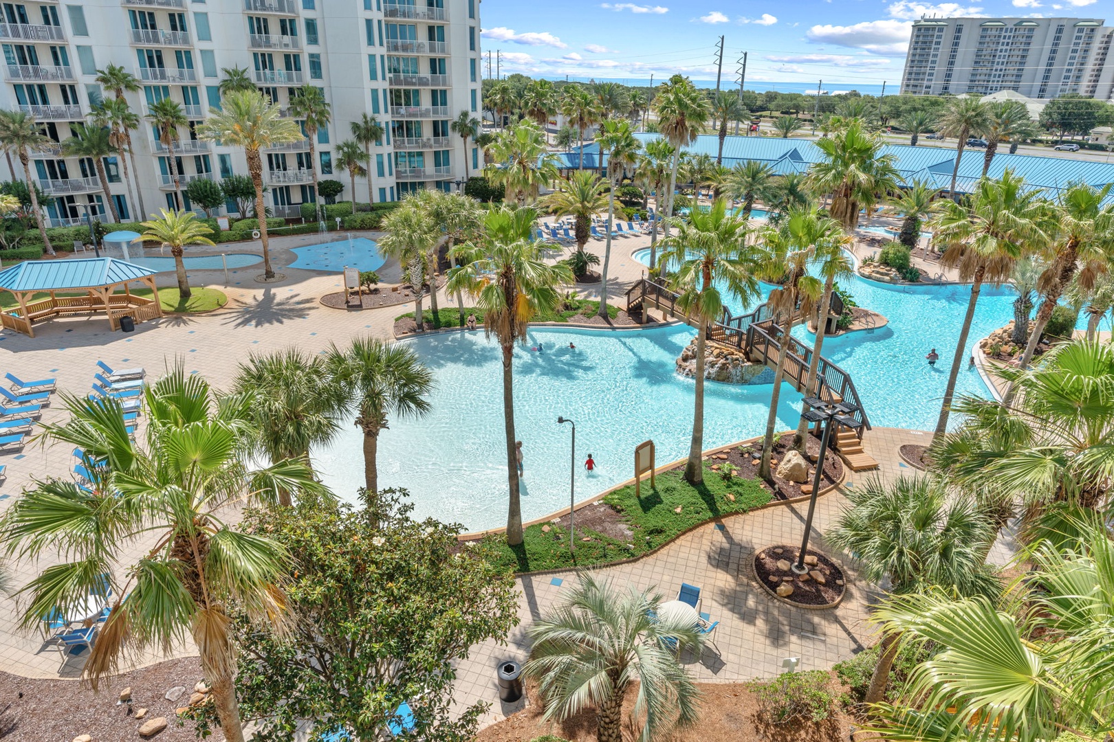 An expansive lagoon-style pool oasis surrounded by swaying palms, lush tropical landscaping, and multi-level resort buildings, with winding pathways and aquatic features throughout.