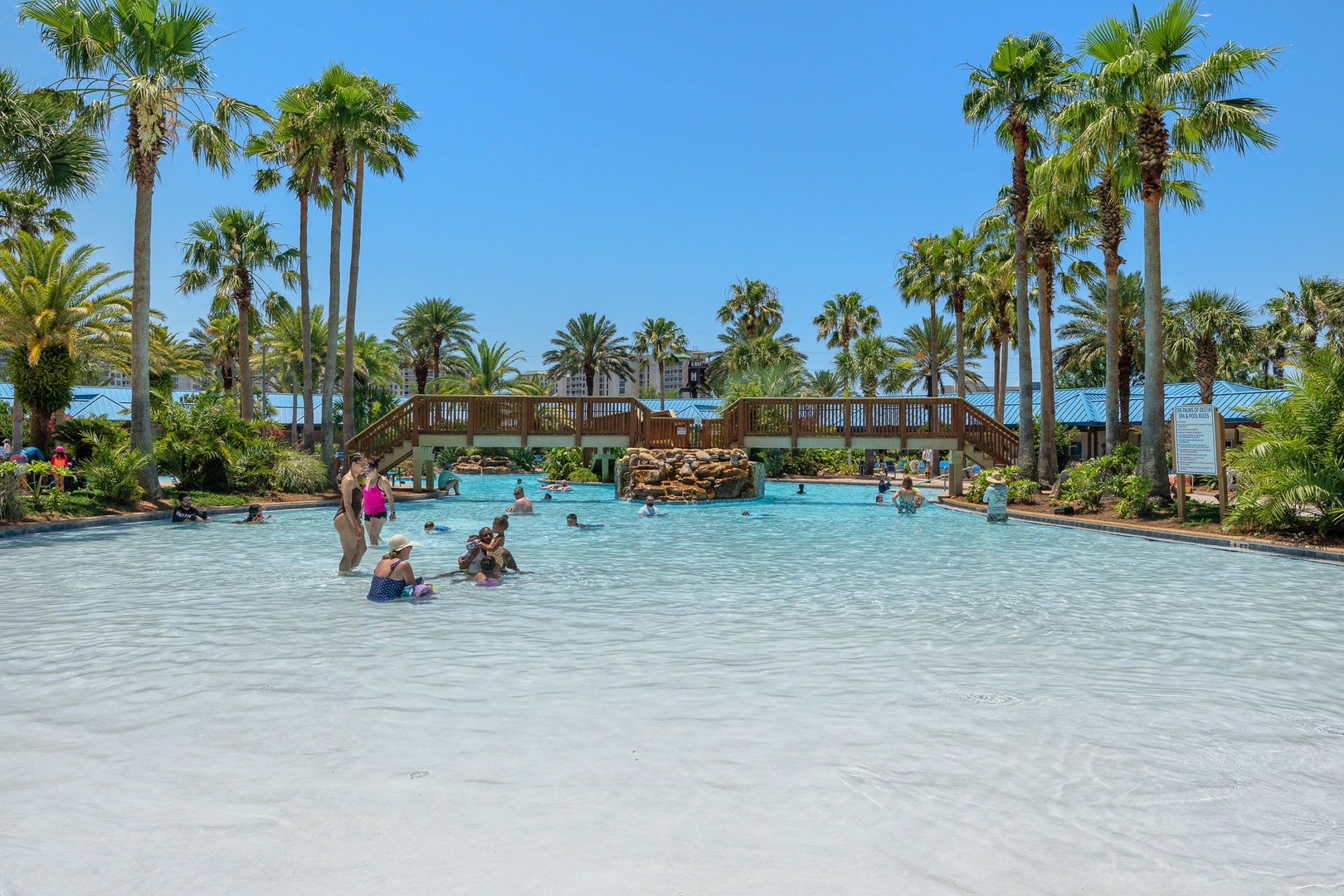 An expansive resort-style pool surrounded by swaying palms and tropical landscaping, featuring a decorative bridge and shallow areas perfect for families to splash and play together.
