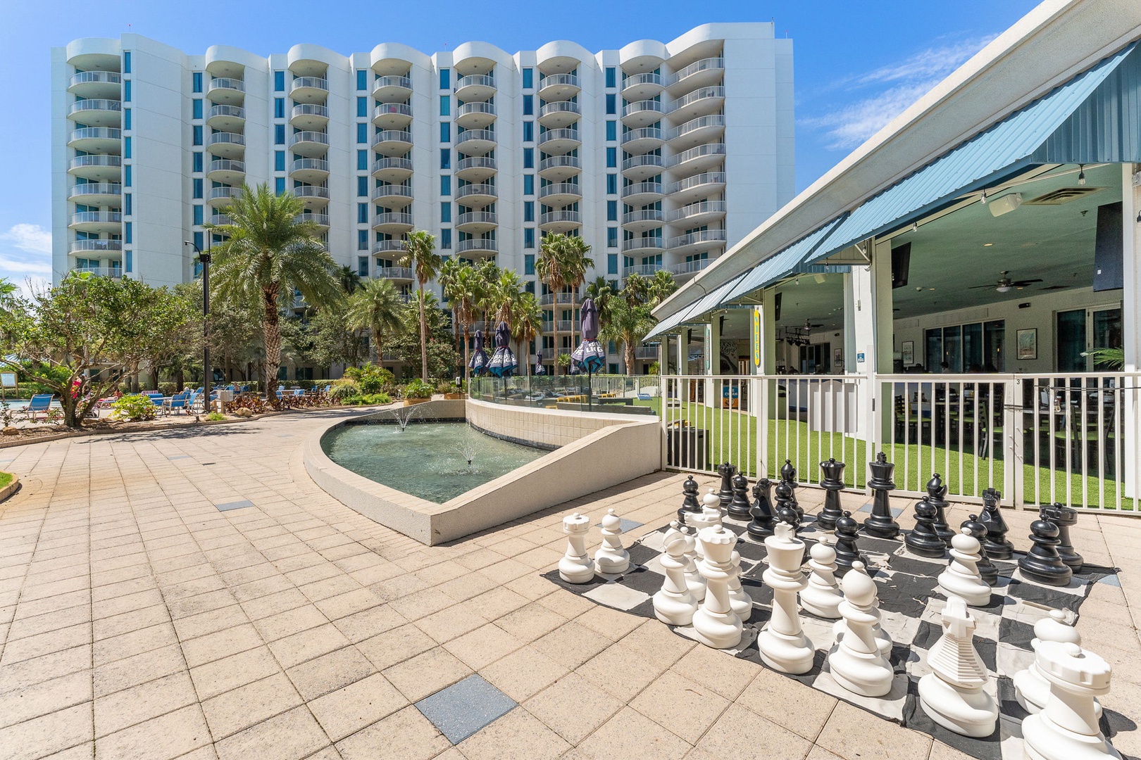 The resort features an outdoor game area with a life-sized chess set, tropical landscaping with palm trees, a decorative fountain, and a modern high-rise building providing coastal accommodations.