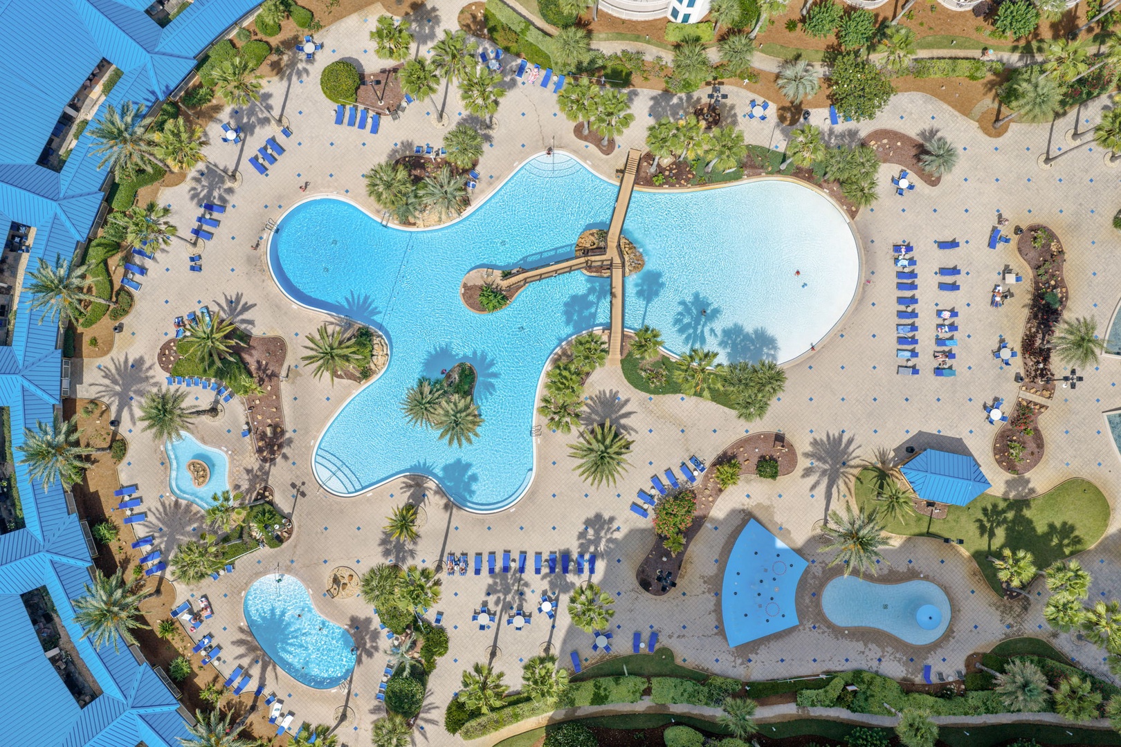 Aerial view of a resort-style aqua park featuring a freeform lagoon pool, water slide, splash zones, tropical palm trees, and blue lounge chairs throughout the landscaped deck area.