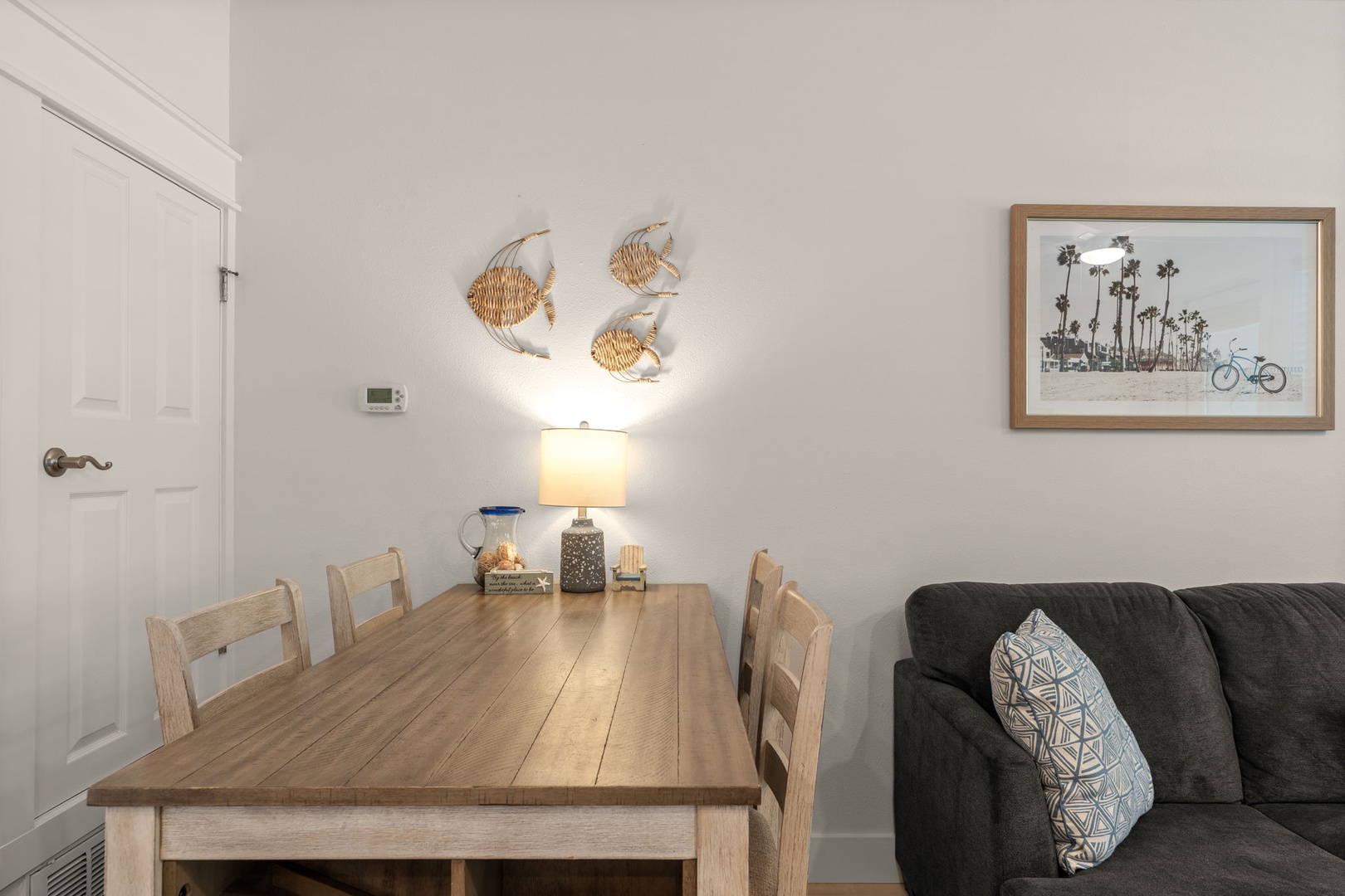Gather around the rustic wooden dining table where morning coffee and easy conversation flow naturally. The coastal artwork and woven wall decor set a relaxed, beach-inspired tone for your stay.