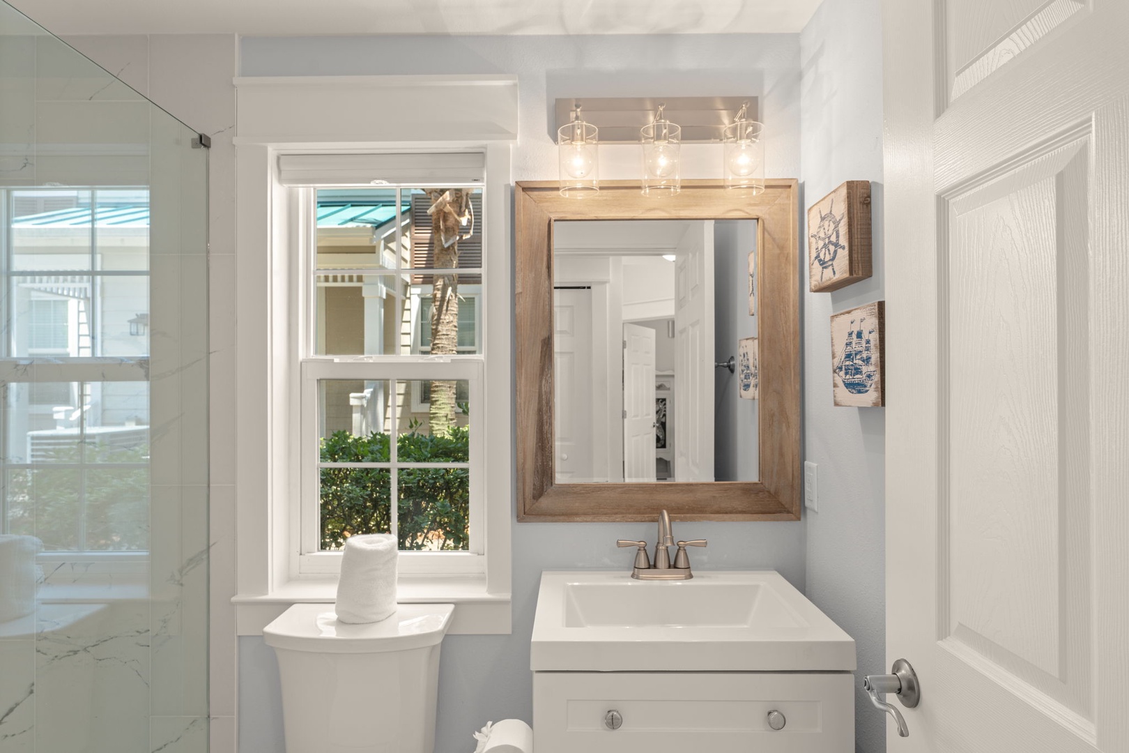 Your bright coastal bathroom features a wood-framed mirror above a crisp white vanity, with nautical touches and natural light streaming through the window to start your mornings feeling refreshed.